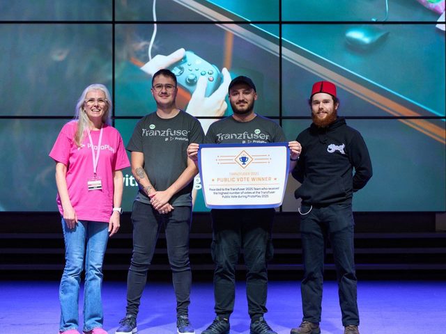 Three Shiverbug Studios team members on the ProtoPlay stage with a host, holding an oversized Tranzfuser 2025 Public Vote Winner certificate
